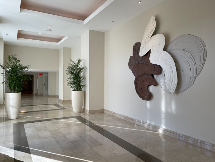 A contemporary abstract artwork by Donald Martiny titled Zimtsterne (2024), displayed in a modern hallway at the Beach Club at the Boca Raton. The piece consists of large, sweeping strokes in shades of white, gray, and brown, appearing as though paint has